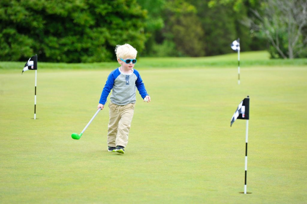 Child on a golf green holding a small golf club and he has sunglasses on.