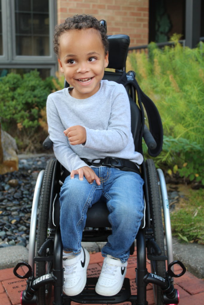 Child smiling while sitting in a wheelchair that is outside near a garden.