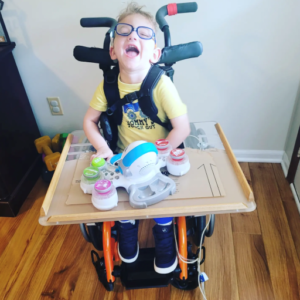 Child smiling at camera while sitting in wheelchair with tray and toy.