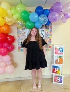 Woman in a black dress, stands in front of balloons and smiles for the camera next to the VIPS sign.