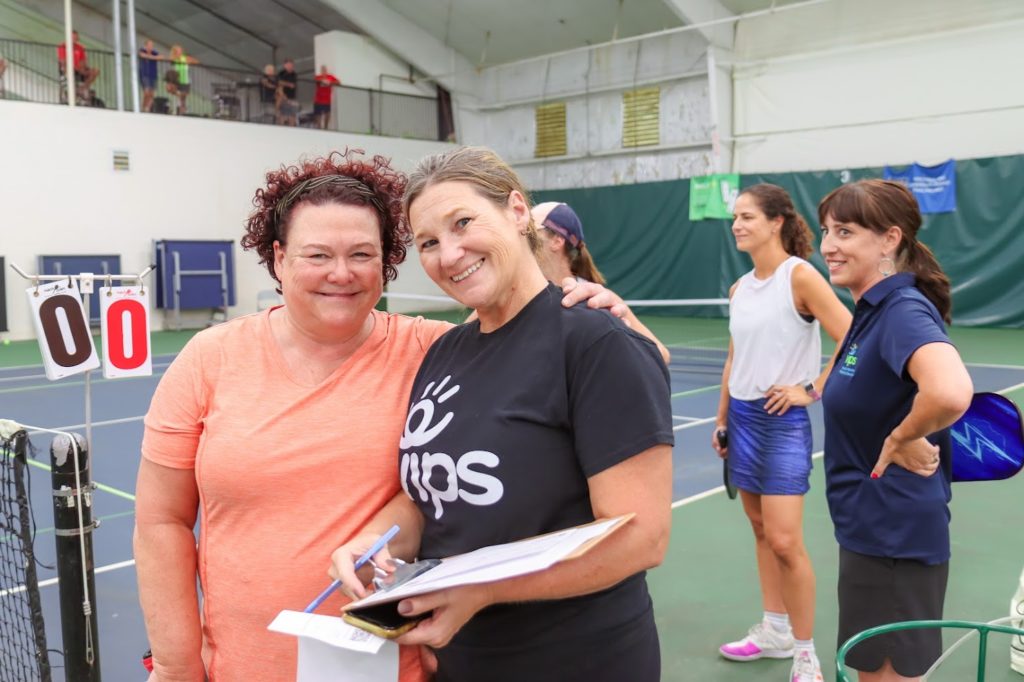 Two women stand at the pickleball court smiling