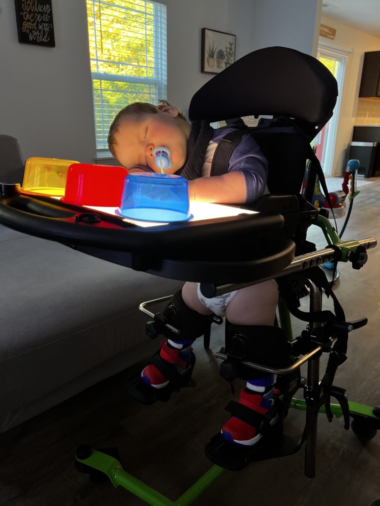 Lucas in his stander with a tray in front of him with light up toys. He is asleep with his head on the tray.