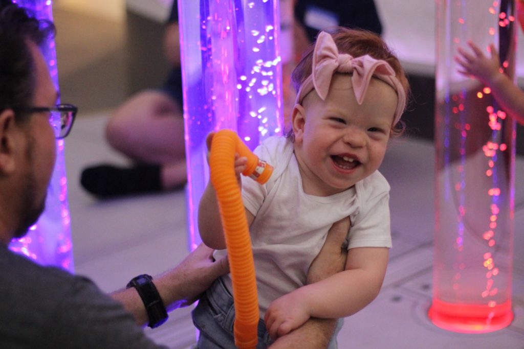 A little girl is smiling while being held by a VIPS teacher. She is in front of a light-up bubble tube.