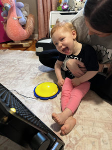 Child sits on the lap of an adult and is smiling while looking forward at assistive technology.