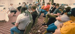 Group of people sitting down some with visual impairment simulator goggles on listening to a story being read by a woman who is seated.