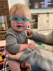 Baby is sitting up with the help of someone, has blue glasses and has a slight smile.