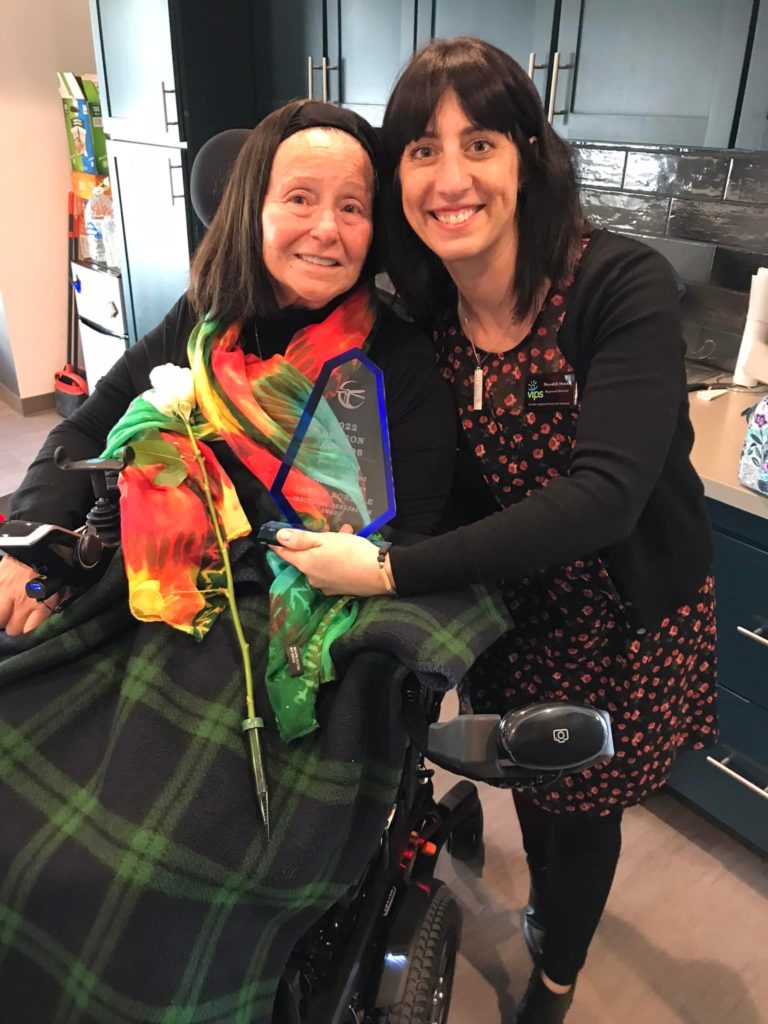 Two women are close to one another smiling. One is in a wheelchair and the other is standing near her holding an award.