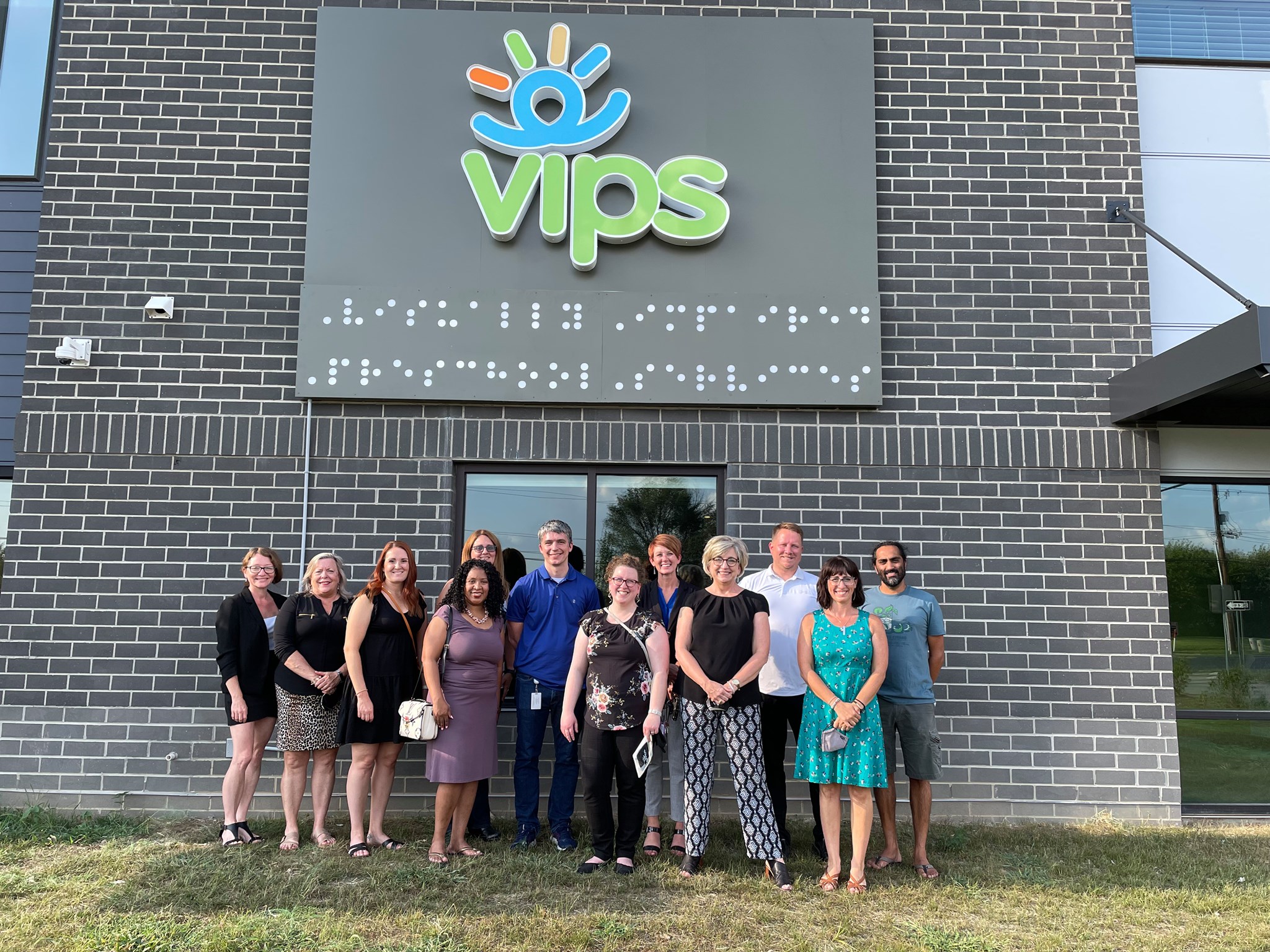 A group of adults are outside standing beneath a sign that says VIPS and braille underneath that reads Visually Impaired Preschool Services