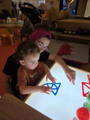 Child sitting with a teacher. The child has a large lightbox in front of them with a blue shape. The child is holding the shape.