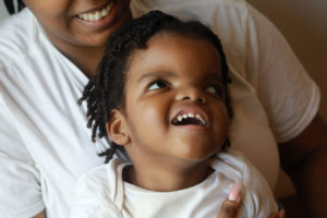 A boy is smiling while looking up and is sitting on the lap of someone who is also smiling. They are both in white shirts.