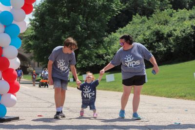 A brother and mom hold hands with a little girl who is smiling as she walks. They are all outside and have shirts that say VIPS on them.