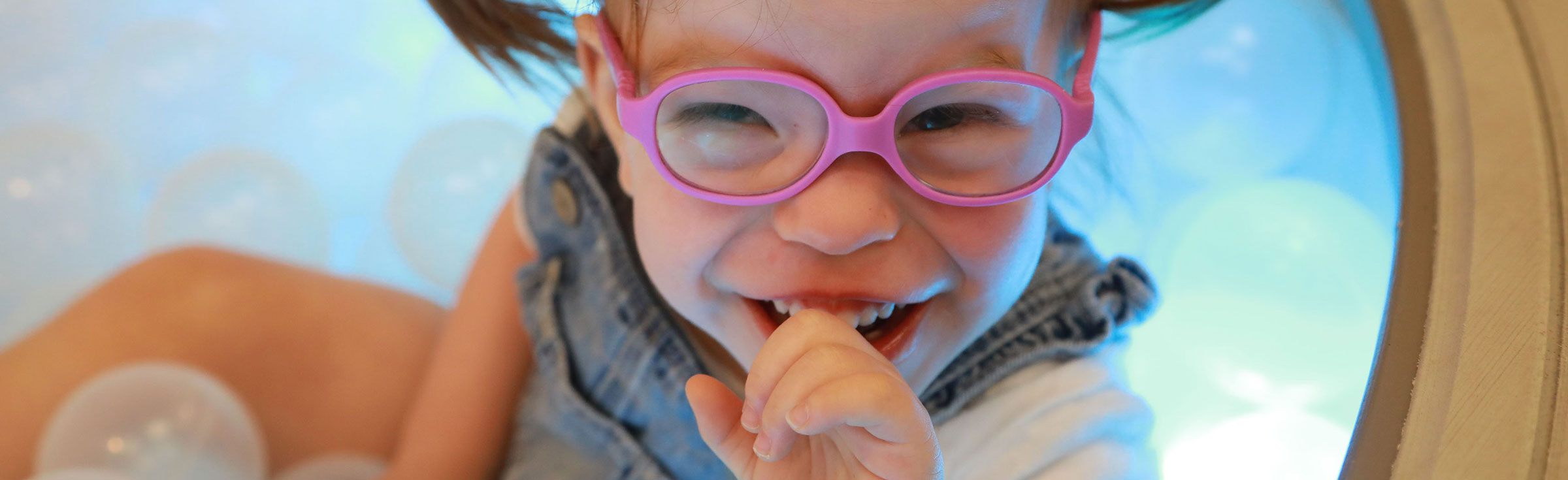 A small girl with pigtails and pink glasses sitting in a sensory ball pit.