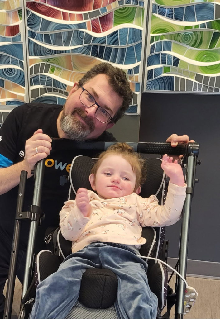 A man with glasses stands behind a young child in a stroller