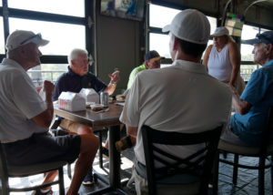 Tom Briggs sitting at a table in the clubhouse talking with his teammates before the scramble begins. Executive Director Diane Nelson stand at the right end of the table talking to the group.