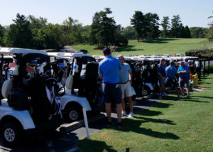 Golf carts lined up before the scramble begins. Several people stand behind the carts talking.