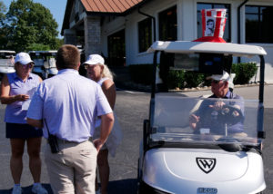 Debbie Baldwin, Josh Griffin, Diane Nelson, and Tom Briggs meeting before the scramble kicks off. Tom Briggs is sitting in his golf cart, adorned with a KFC bucket and the image of the Colonel has been replaced with a likeness of Tom.