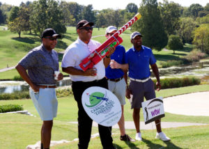 Four men pose for a photo in front of the Cannonball Experience sign. One golfer holds the cannon with both hands. The cannon shoots a golf ball across the course. Golfers can choose this instead of teeing off at the hole.