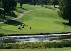 Two men and two golf carts on the fairway against a hill. In the foreground, stream runs through the fairway.