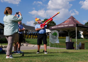 A man wearing a hat that looks like a roasted chicken shoots a golf ball from the air cannon.