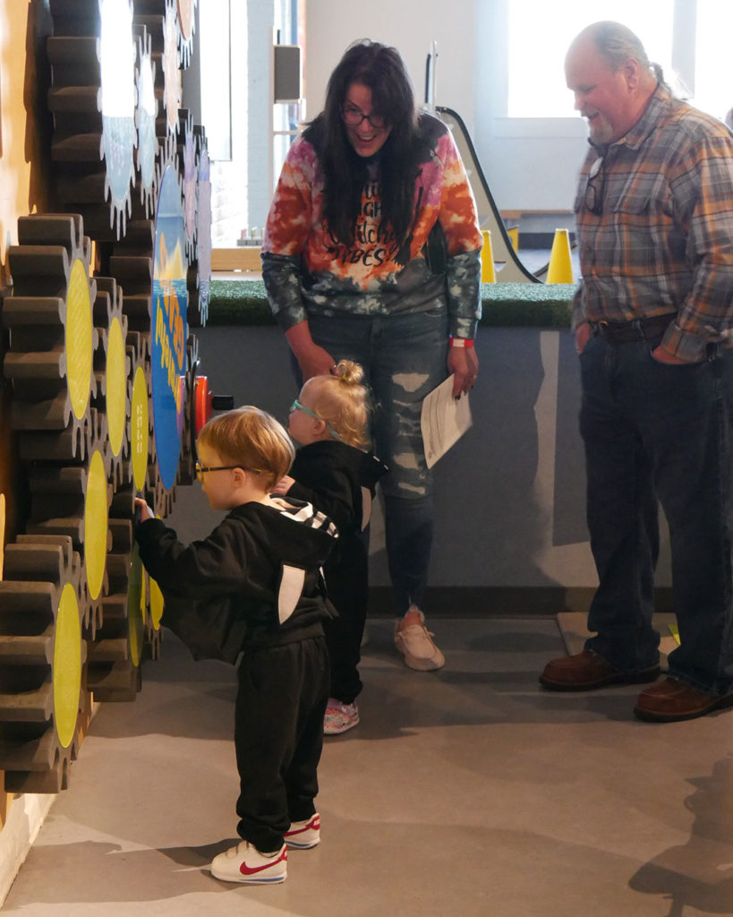 A small boy and a girl turn giant gears mounted to the wall at the Kentucky Science Center while their grandparents smile and encourage them.