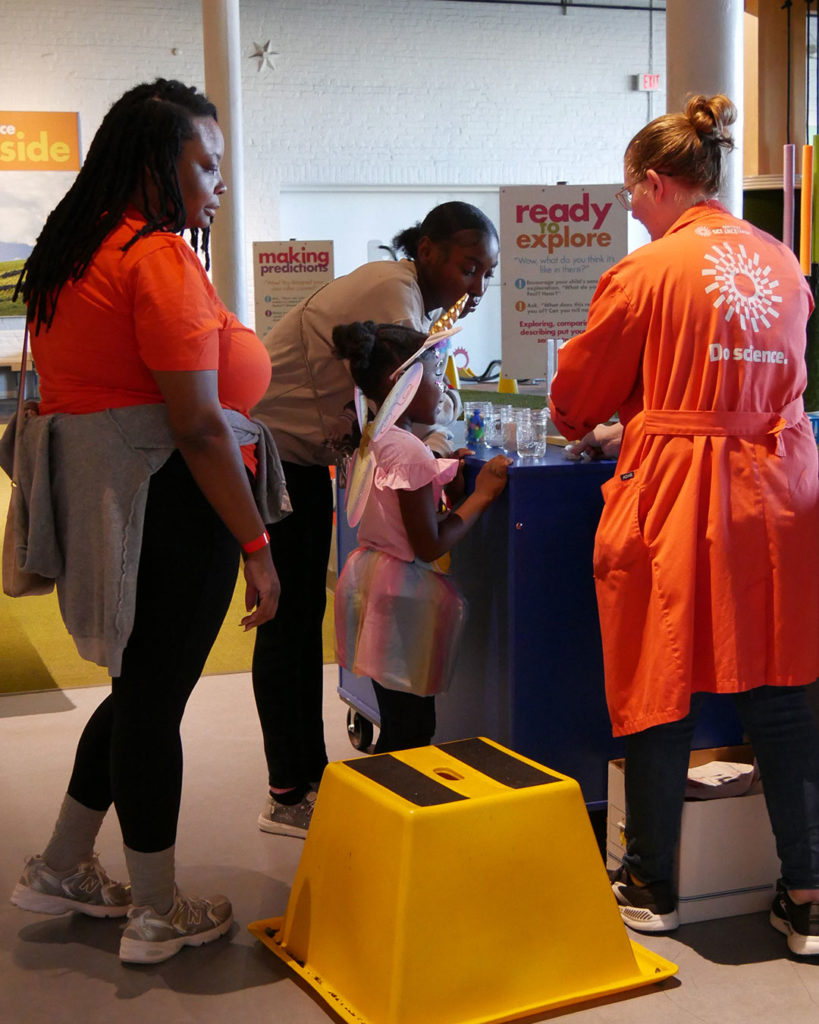 A young girl wearing a unicorn horn and fairy wings participates in a science experiment with a member of the Ky Science Center staff while her family watches.