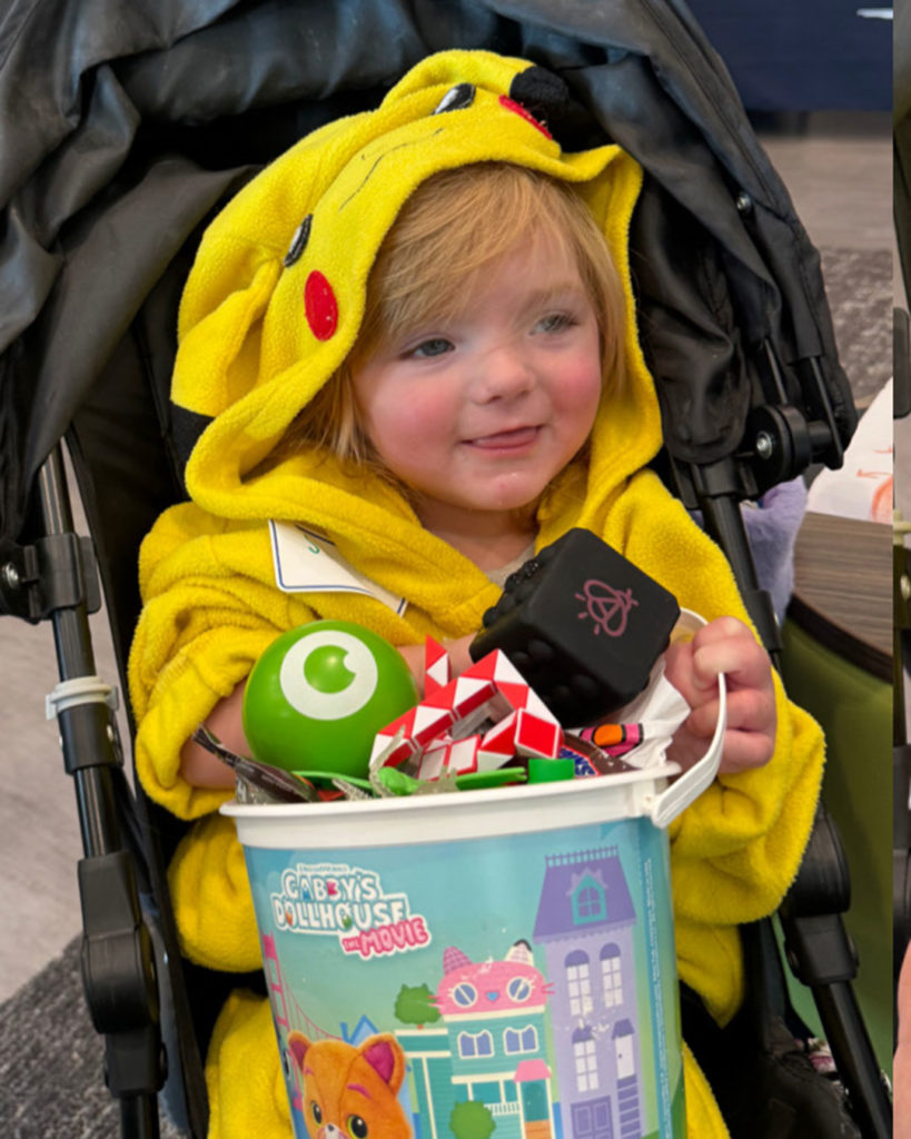 A small girl wearing a Pokémon costume sits in a stroller holding a bucket of toys.