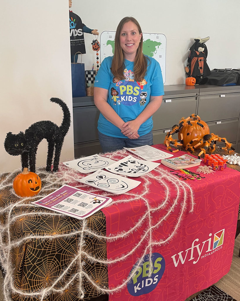 A woman representing WFYI/PBS Kids poses for a photo behind their table at the FRC, which is decorated with pumpkins, spiders, and a black cat.