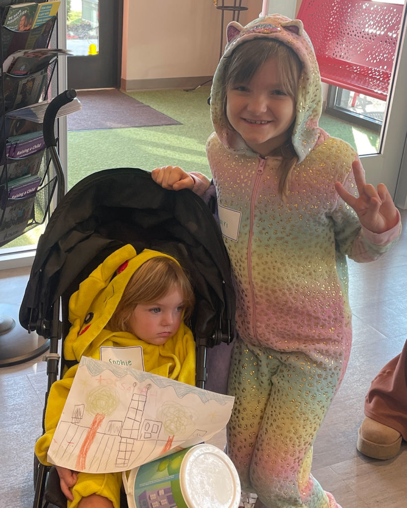 A small girl wearing a Pokémon costume sits in a stroller. A young girl wearing a rainbow costume with cat ears stands beside her, flashing a peace symbol.