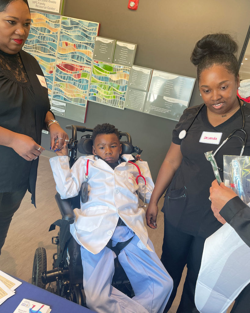 A boy wearing a doctor's costume sits in a mobility chair at the Oral Health Division of the Indiana Department of Health table.