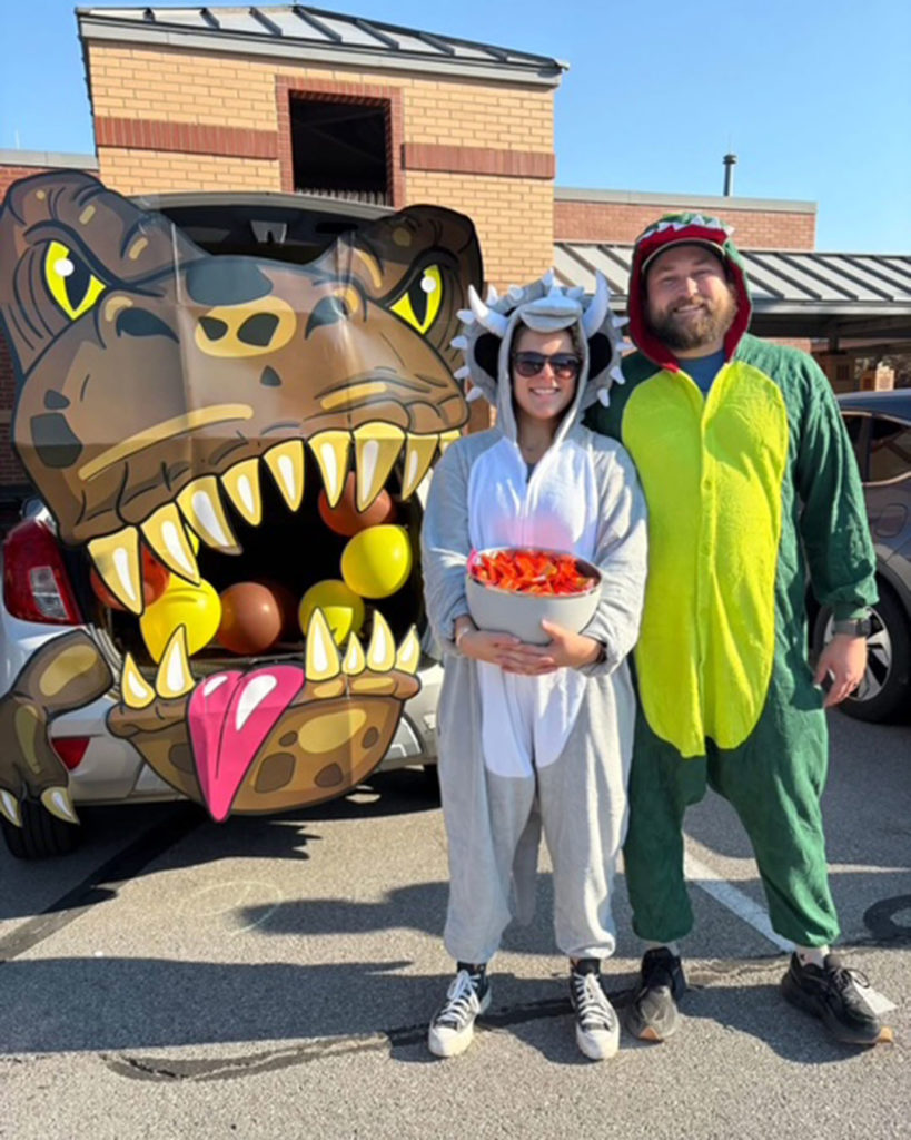 A woman and a man in dinosaur onesies pose for a photo in front of a large T Rex cutout.