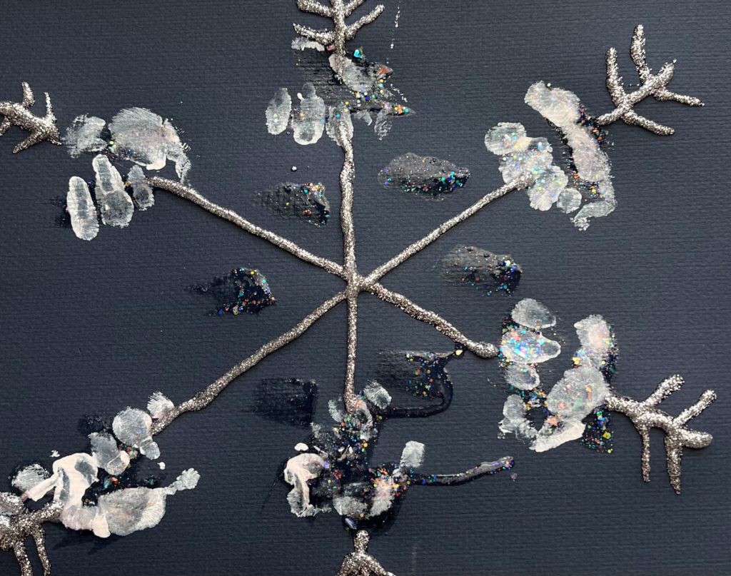 A six point child-made snowflake created with glitter glue and inward-pointing chevrons on the ends on black construction paper. The spines are decorated with handprints in white ink.