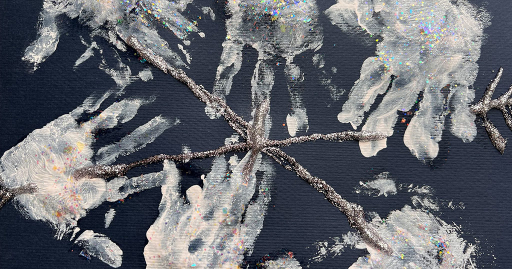 A six point child-made snowflake created with glitter glue and inward-pointing chevrons on the ends on black construction paper. The spines are decorated with handprints in white ink.