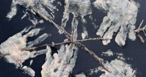 A six point child-made snowflake created with glitter glue and inward-pointing chevrons on the ends on black construction paper. The spines are decorated with handprints in white ink.