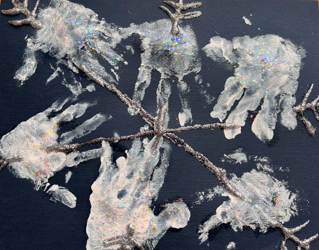A six point child-made snowflake created with glitter glue and inward-pointing chevrons on the ends on black construction paper. The spines are decorated with handprints in white ink.