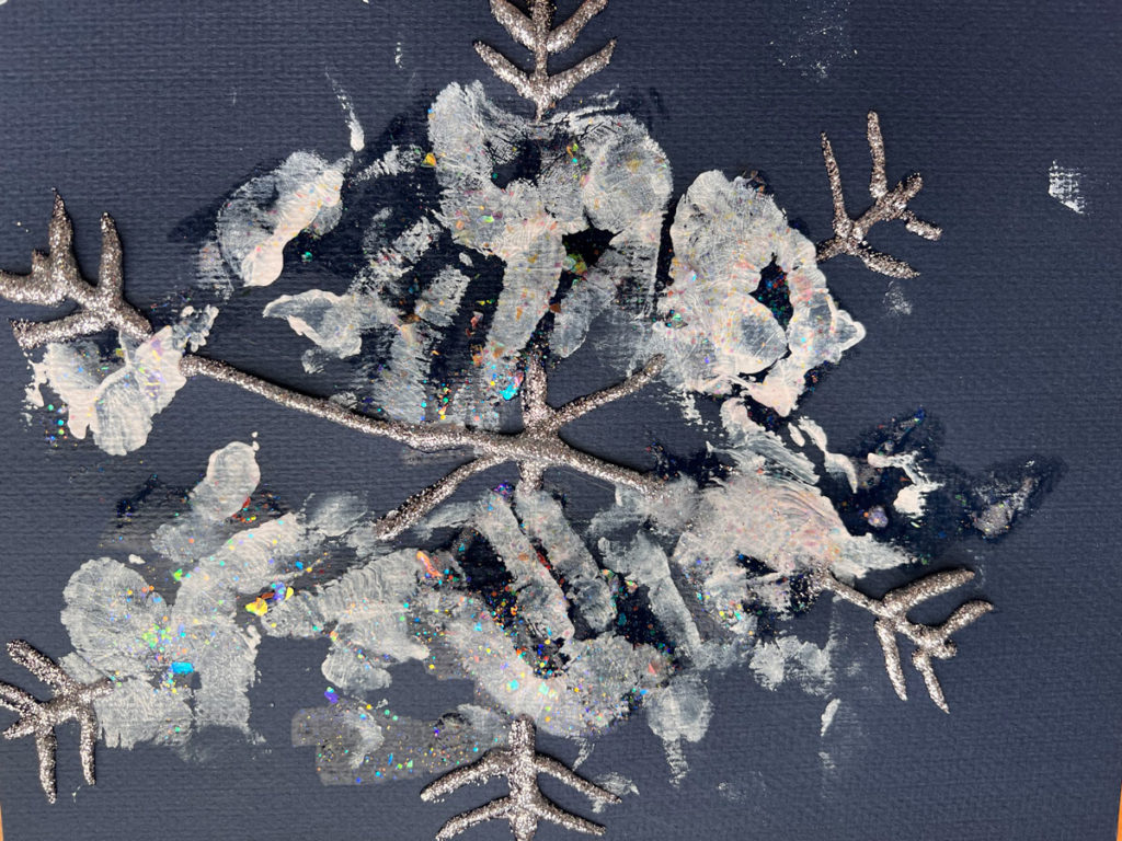 A six point child-made snowflake created with glitter glue and inward-pointing chevrons on the ends on black construction paper. The spines are decorated with handprints in white ink.