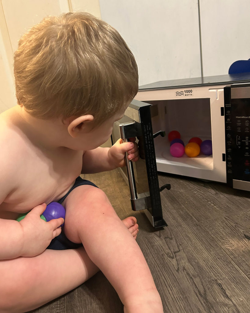 Phoenix sits on the floor and is placing multicolored plastic balls in his microwave.