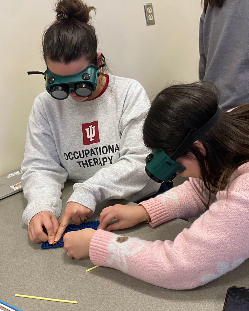 Two female OT students attempt to use a sensory activity board while wearing simulation goggles.