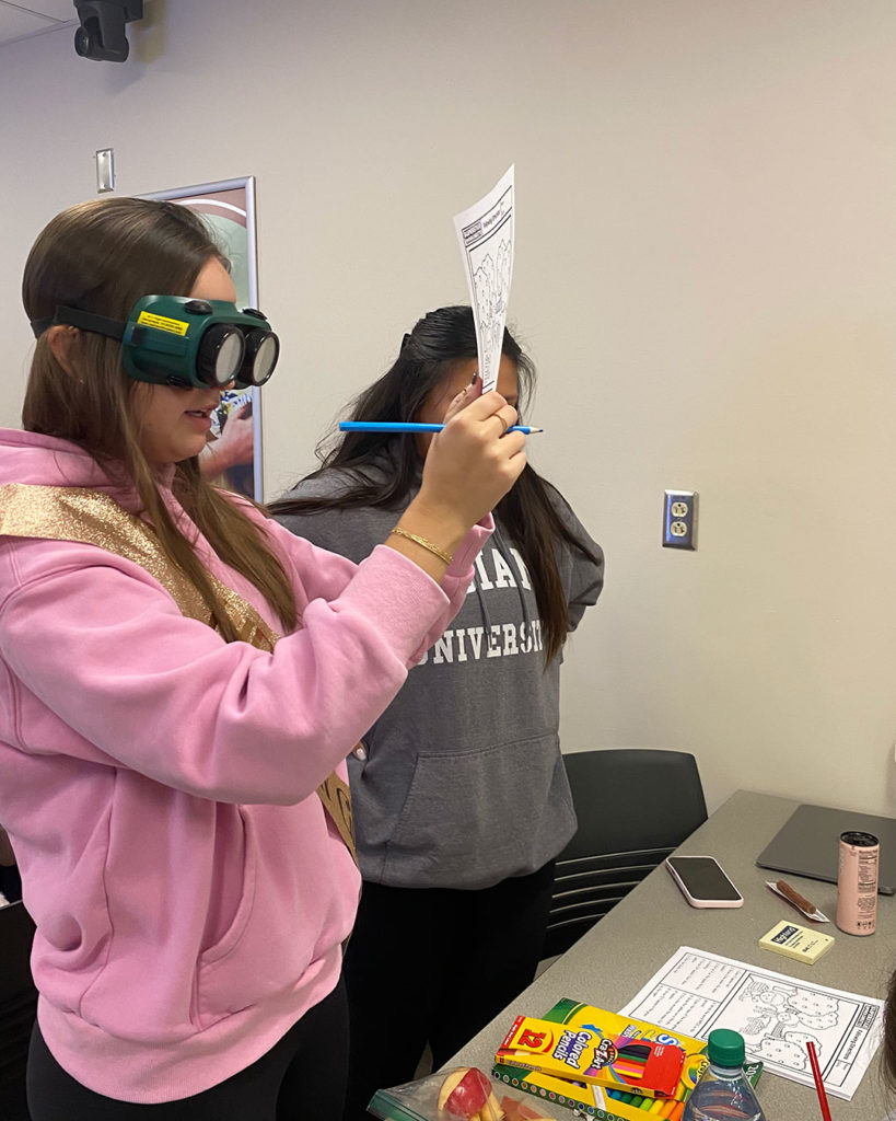 A female OT student is holding up a coloring worksheet and trying to read it while wearing simulation goggles.