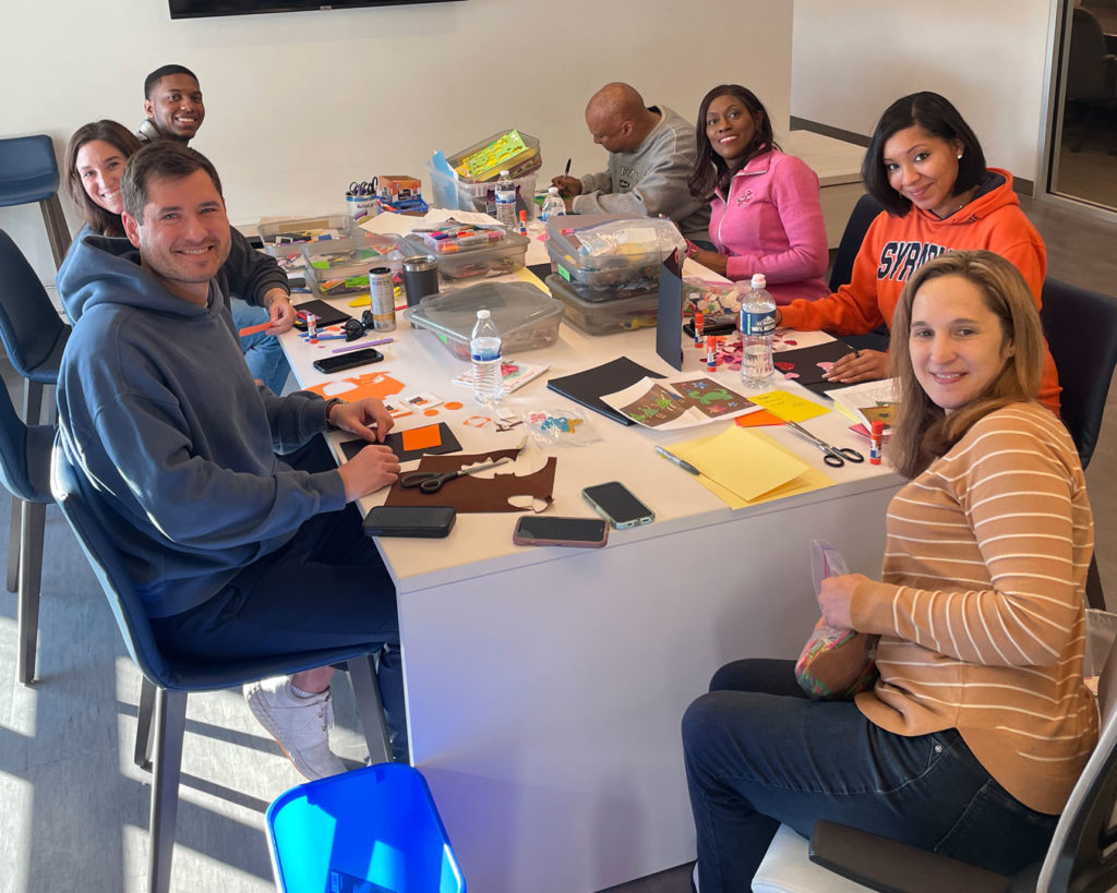Volunteers from the NCAA PR team turn and pose for a photo while sitting at the FRC work table, making cards.