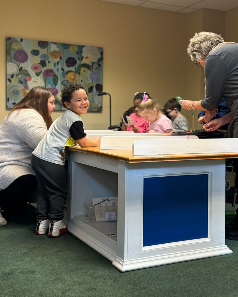A small boy turns and smiles for the camera while playing in the banking center with his classmates.