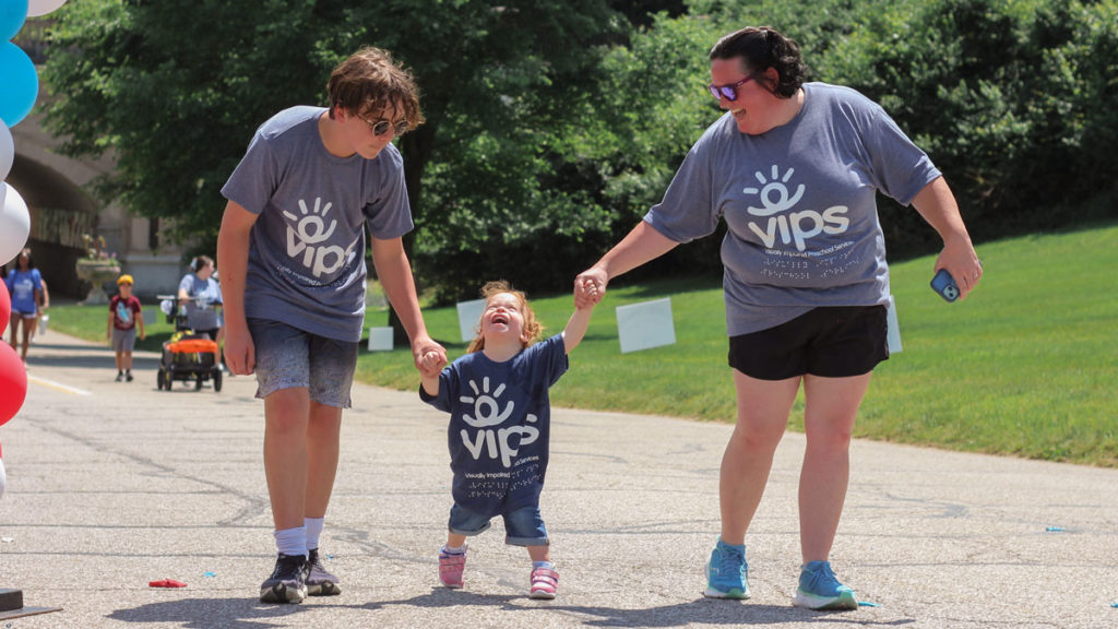A brother and mom hold hands with a little girl who is smiling as she walks. They are all outside and have shirts that say VIPS on them.