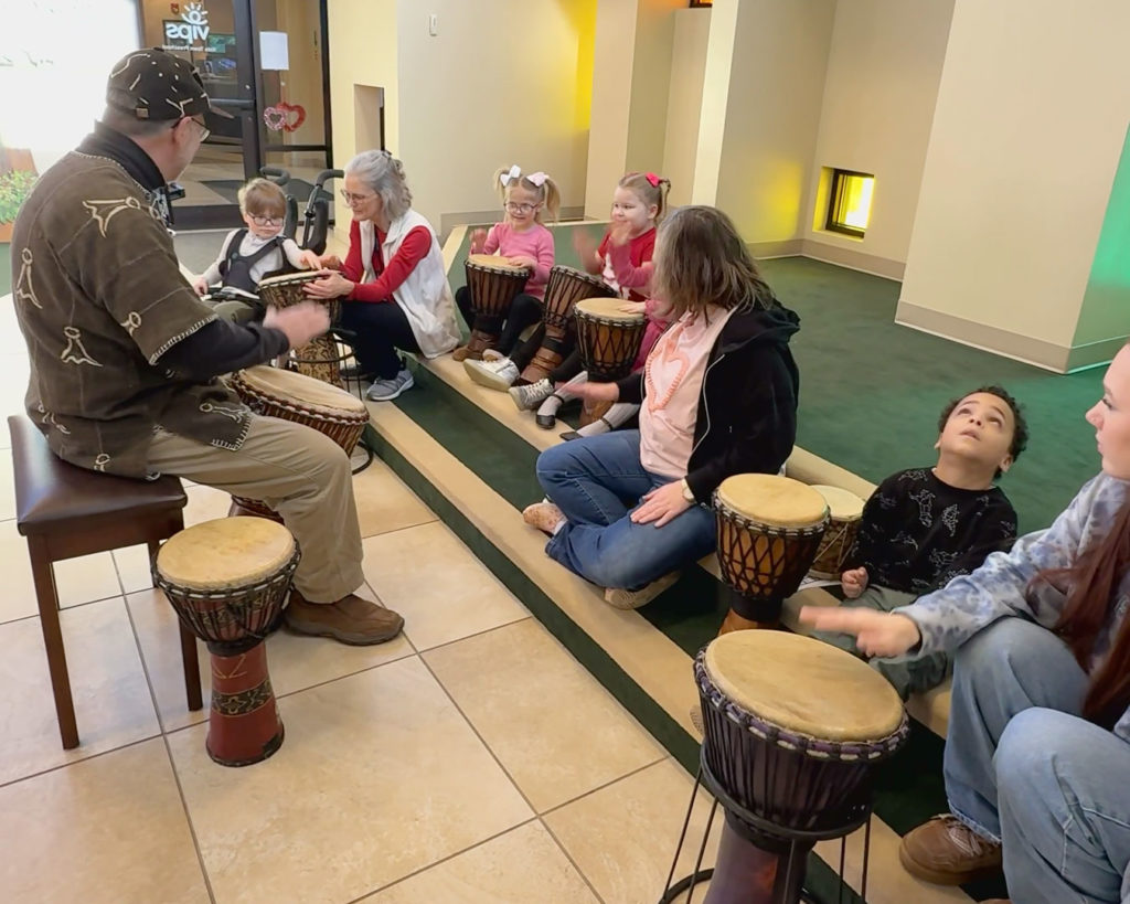 The kids sit on the steps in Kids Town and play drums with Gregory.