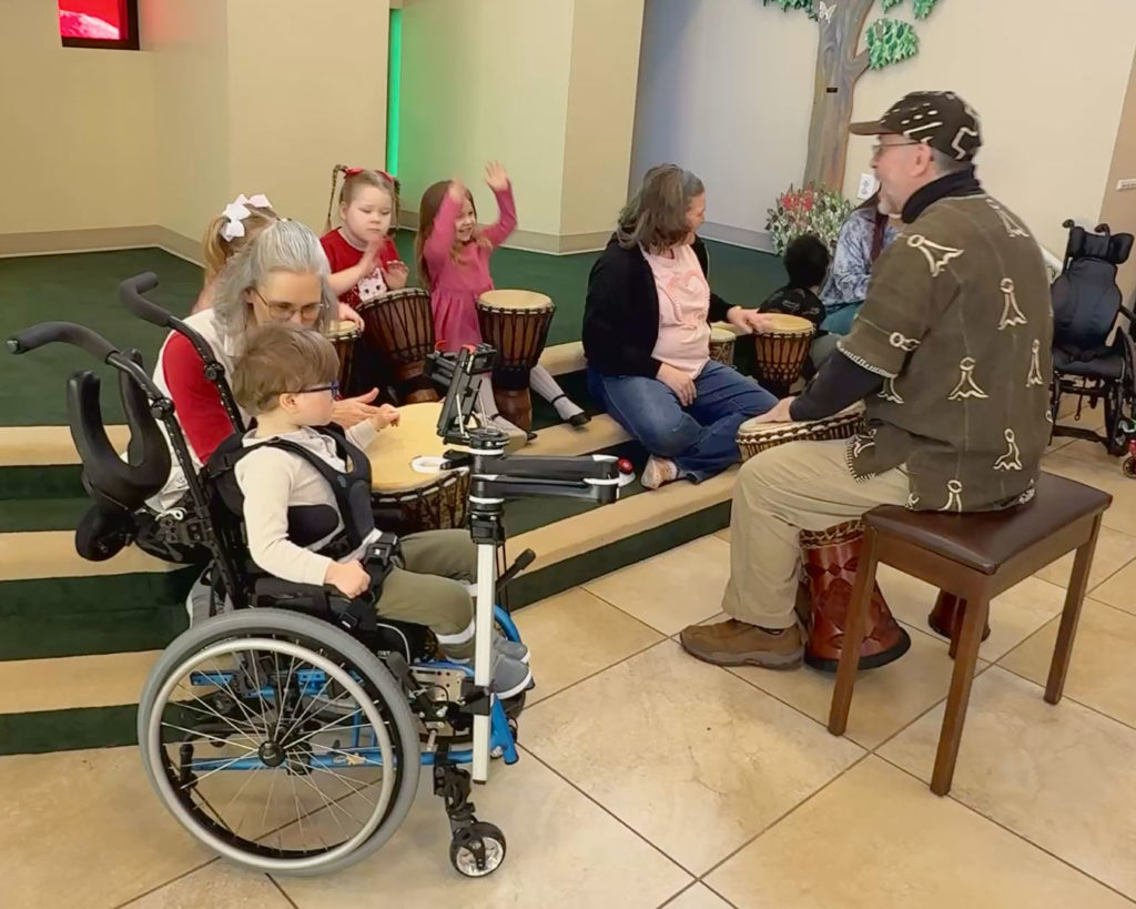 The kids sit on the steps in Kids Town and play drums with Gregory.