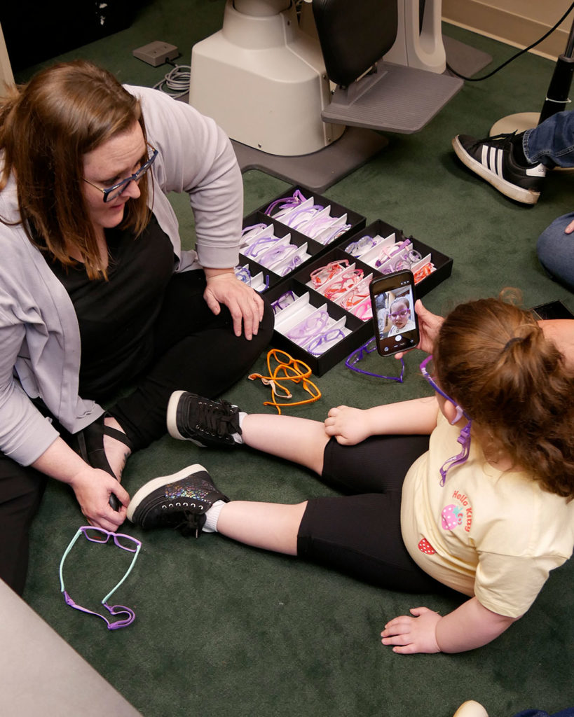 Megan and a small girl sit on the floor with a tray of sample frames. The girl admires the new frames in a phone screen.