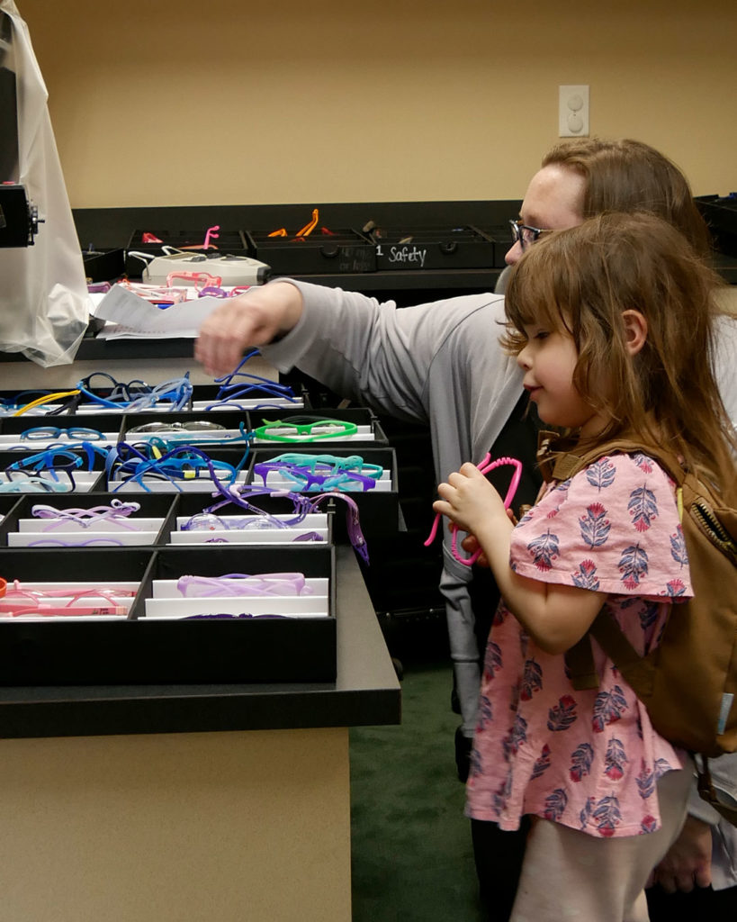 Megan and small girl look at trays of frames and pick some out to try on.