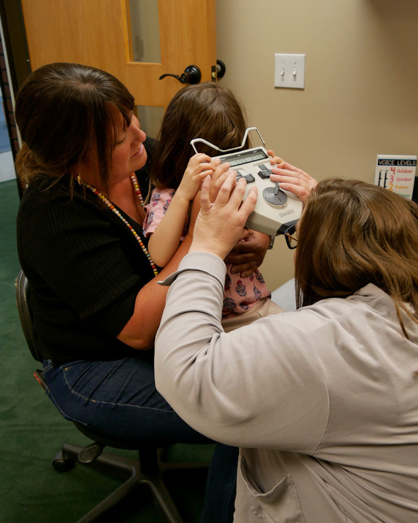 A small girl sits in Ms. Ashley's lap while Megan measures her eyes.