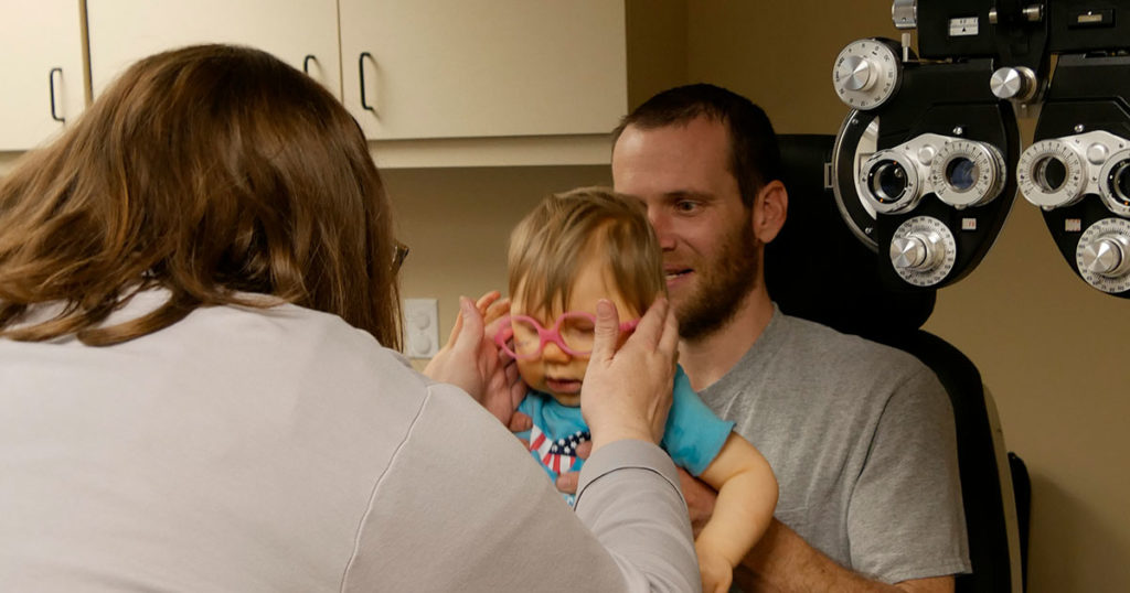 A small boy sits in his father's lap while Megan checks a pair of frames for fit.