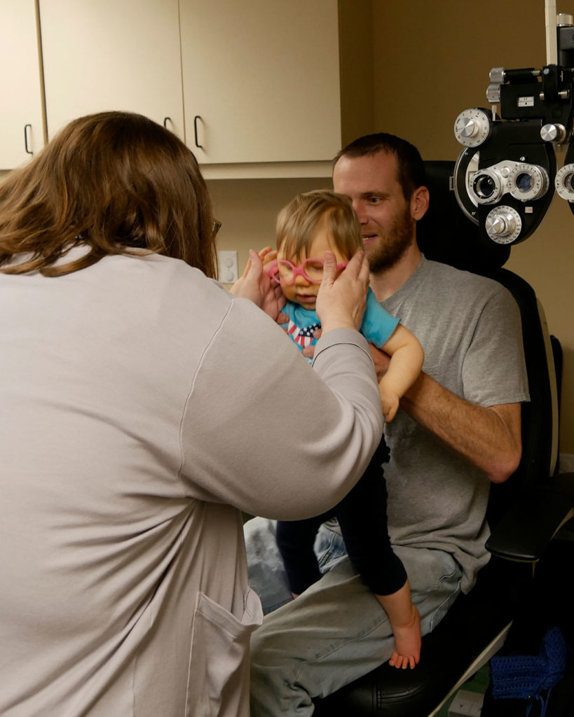 A small boy sits in his father's lap while Megan checks a pair of frames for fit.