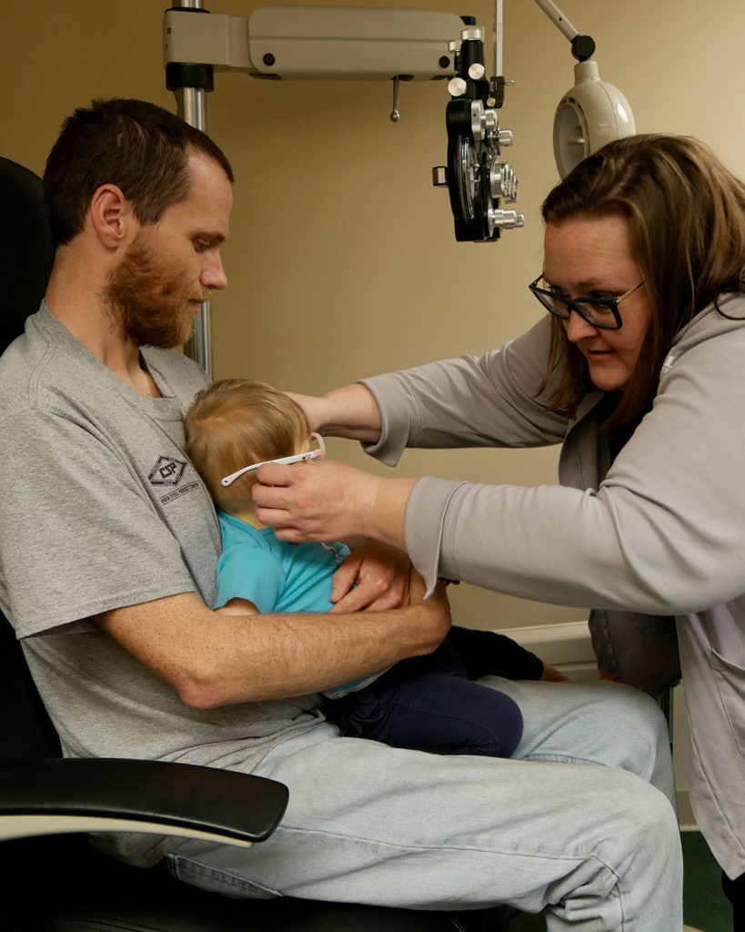 A small boy sits in his father's lap while Megan checks a pair of frames for fit.