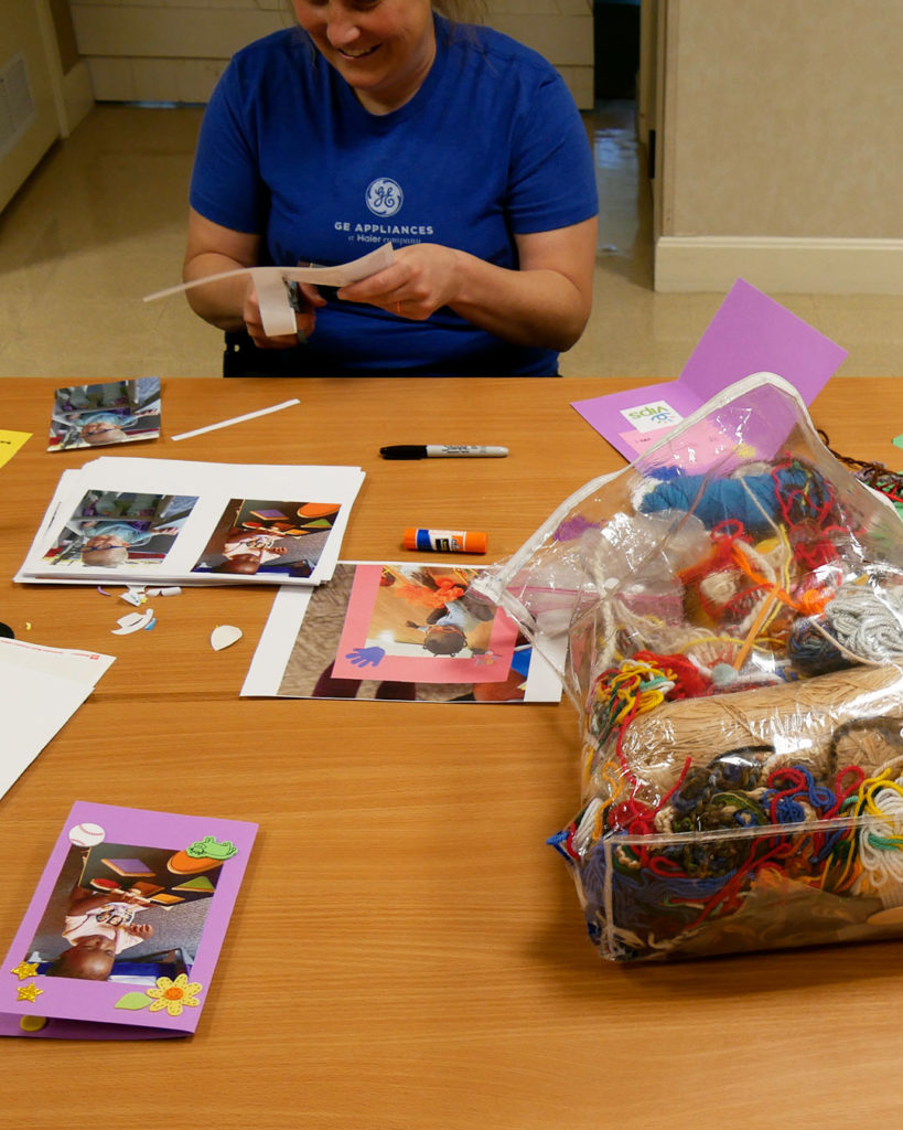 A GE volunteer cuts out photos of kids, and a bag of craft supplies sits in the middle of the table.